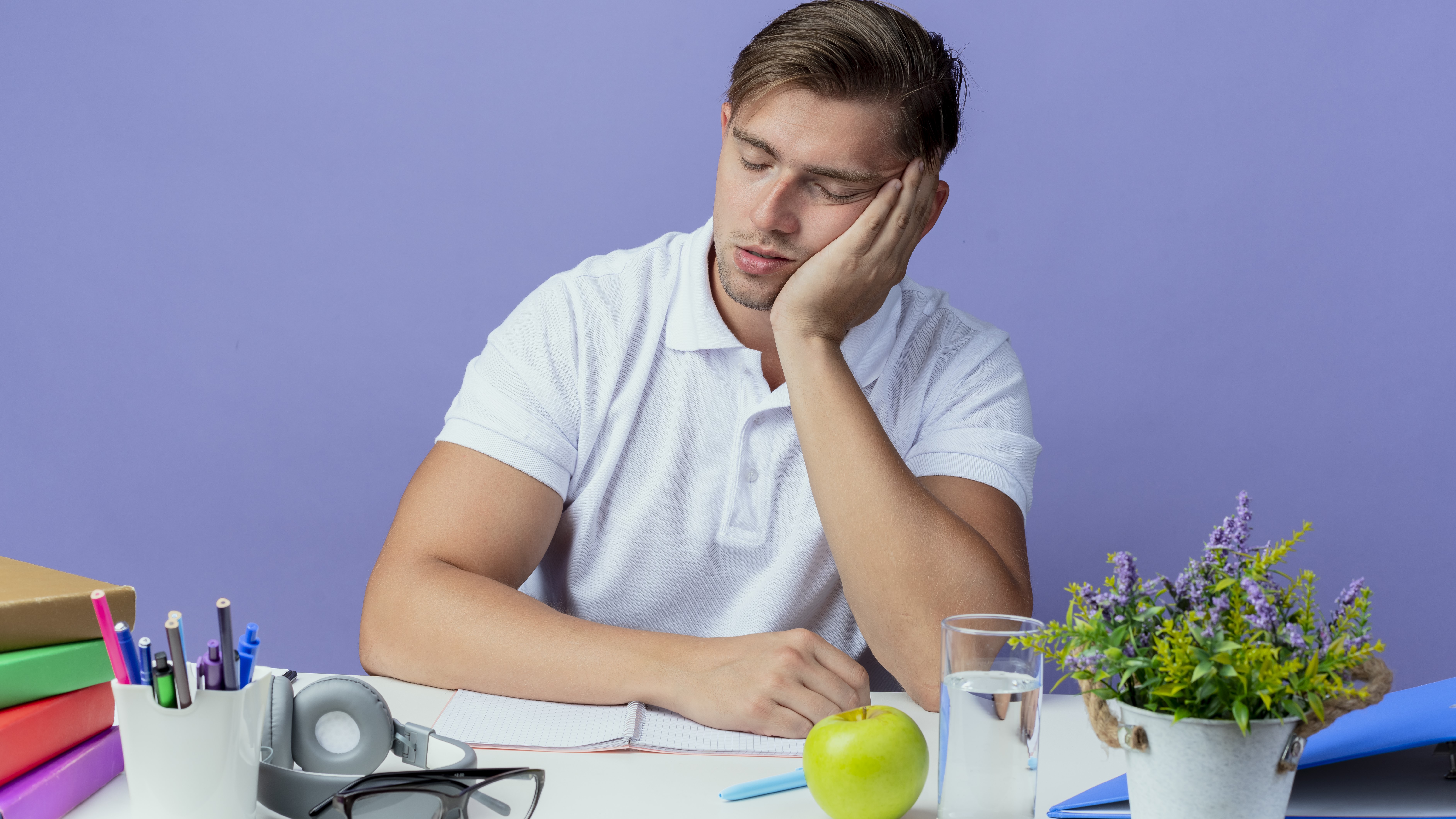 С закрытыми глазами молодой красивый студент-мужчина сидит за столом и спит